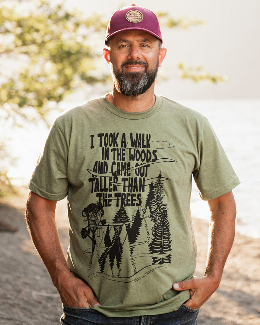 Man wearing a green t-shirt with a graphic design and text, standing outdoors by a body of water.