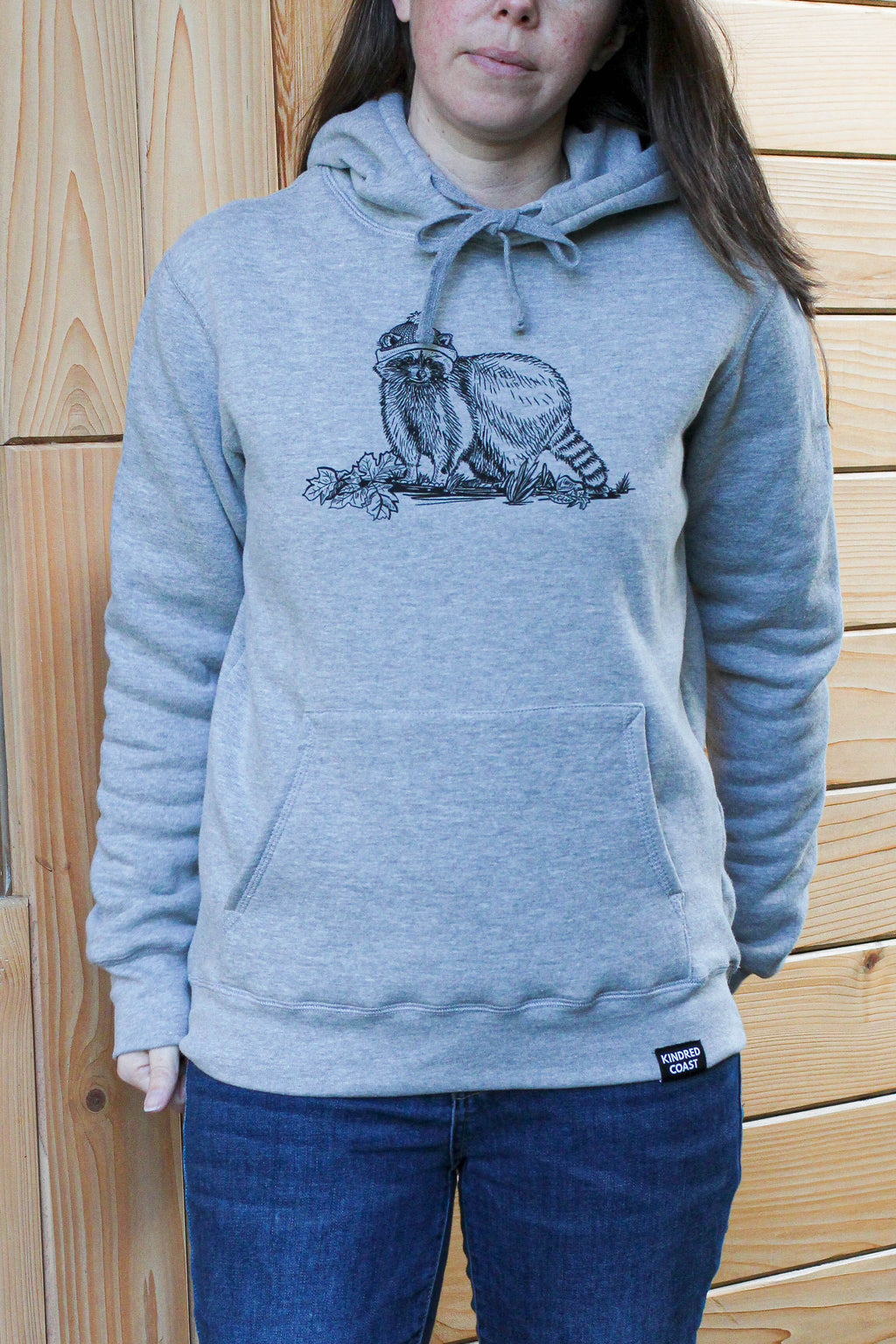 Woman wearing a gray hoodie with a graphic design of a raccoon in front of a wooden wall