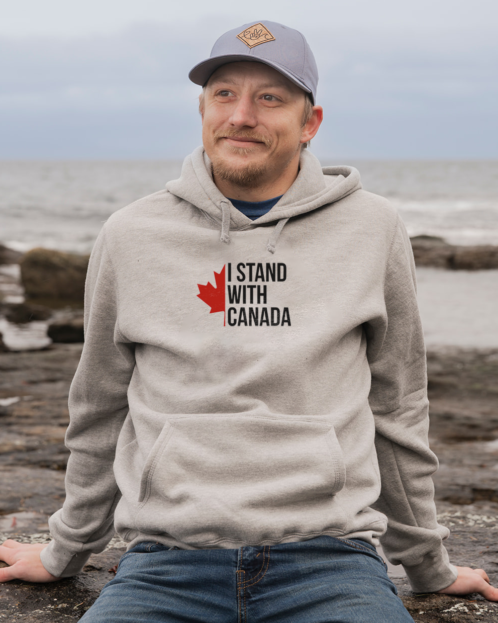 Man wearing a hoodie with 'I Stand With Canada' text and maple leaf graphic by the ocean.