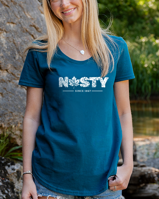 A smiling blonde woman stands outdoors next to a large rock and a stream, wearing a teal t-shirt with 'NASTY' printed in white across the chest, paired with denim shorts.