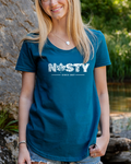 A smiling blonde woman stands outdoors next to a large rock and a stream, wearing a teal t-shirt with 'NASTY' printed in white across the chest, paired with denim shorts.