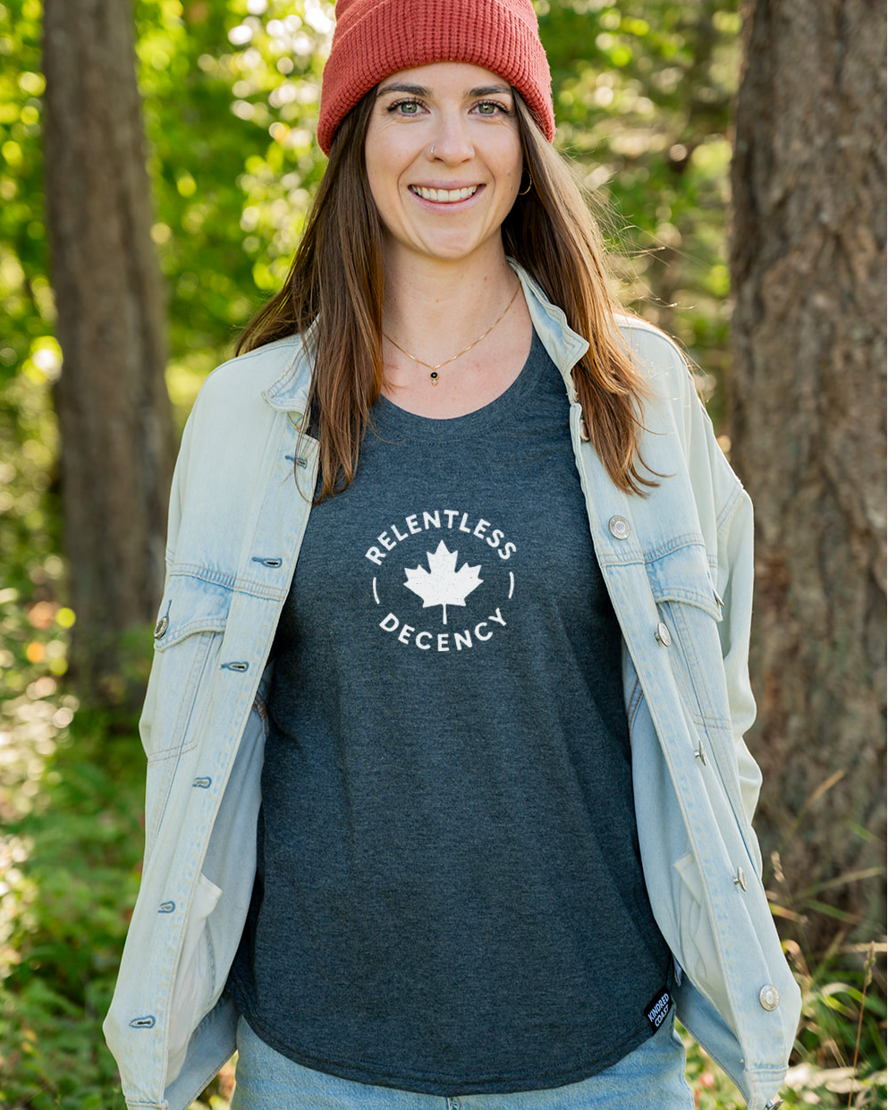 Woman wearing a red beanie and a blue shirt with a logo, standing in a forest.