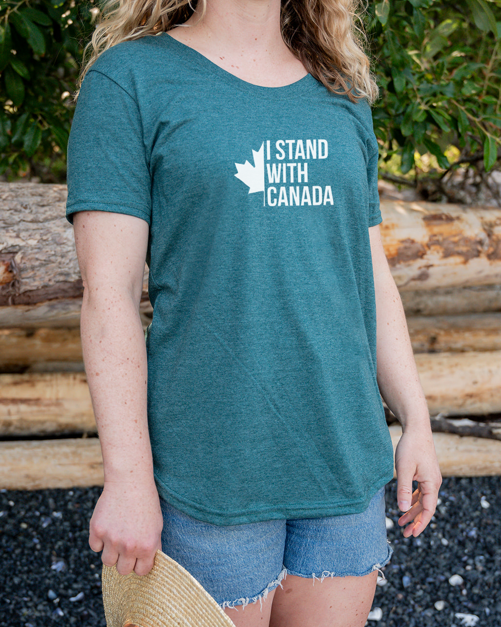 Person wearing a teal t-shirt with a logo and text, standing outdoors with logs and foliage in the background.