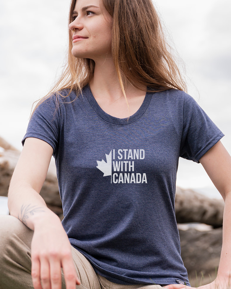 Woman wearing a 'I Stand with Canada' t-shirt sitting on grass with a scenic background